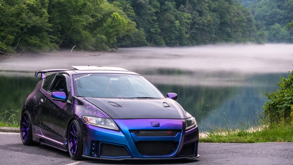 Team Hi Def A purple and blue exotic car, likely an import tuner prized by the local car club, is parked near a serene lake with mist hovering above the water. Lush green forest surrounds the area, creating a calm and scenic backdrop.