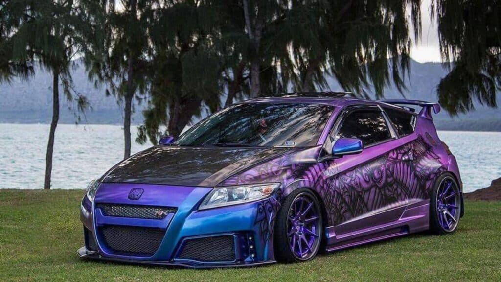 Team Hi Def A custom import tuner with a black, blue, and purple paint job showcases intricate tribal designs. Parked on grass near water with trees in the backdrop, this performance car boasts modified bodywork and striking purple rims.