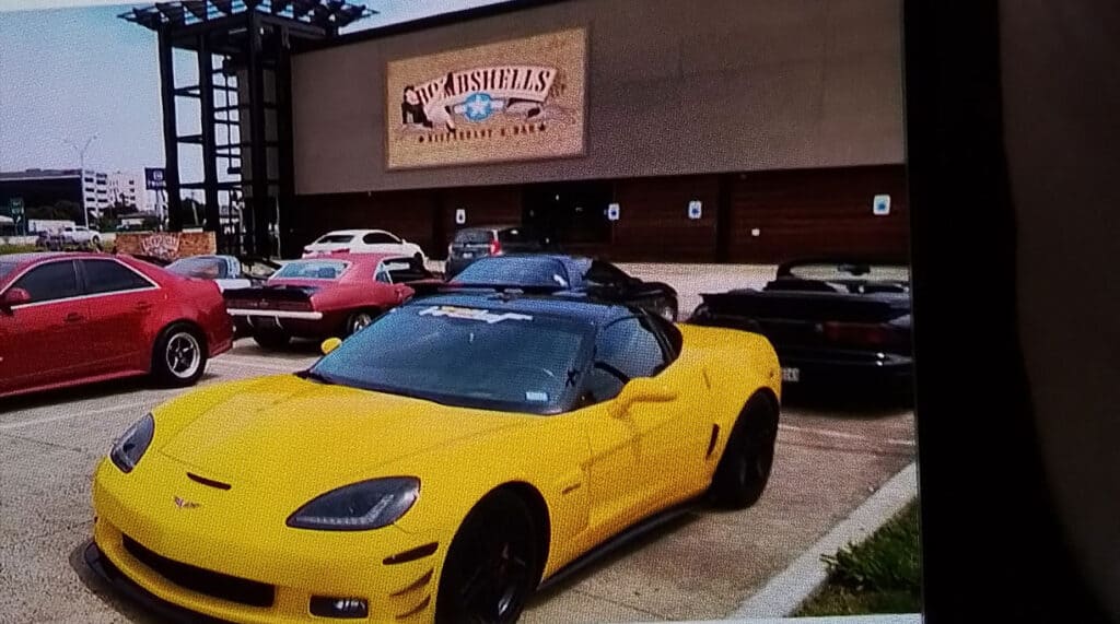 Team Hi Def A yellow muscle car is parked in front of a building with a sign that reads "Texas Bullsh!tters Country Club." Other vehicles are nearby, including red and black ones. The scene unfolds in a parking lot, hinting at an impromptu car club gathering.