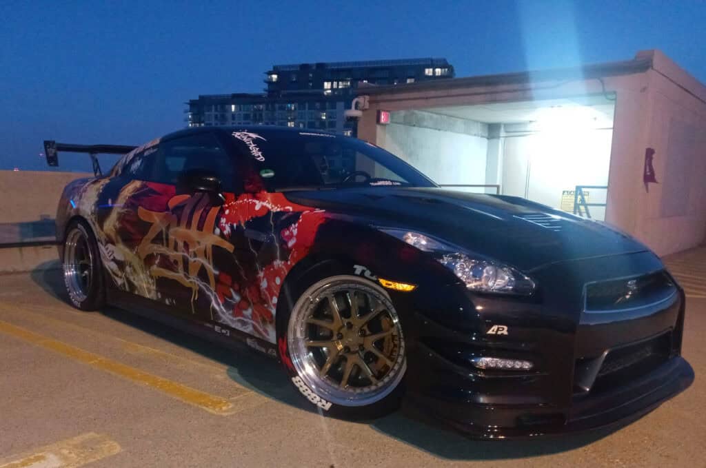 Team Hi Def A sleek, modified sports car boasting colorful graffiti-style artwork on the side is parked on a rooftop at dusk. This JDM-inspired creation features a prominent spoiler and custom wheels, with city buildings shimmering in the background.