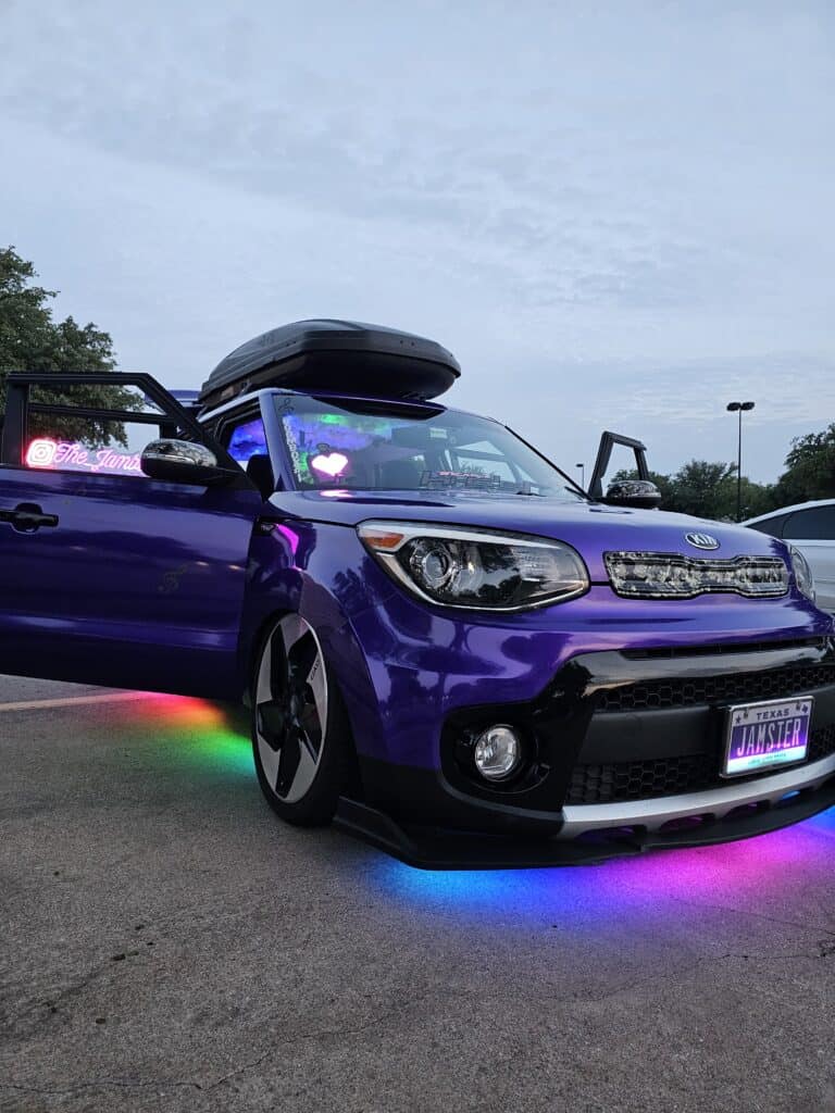 Team Hi Def A vibrant purple import tuner with its doors open is parked on a concrete surface. It features custom underglow lighting in various colors. The license plate reads "JAMSJER," and the interior emits a soft glow. Trees and a cloudy sky are visible in the background.