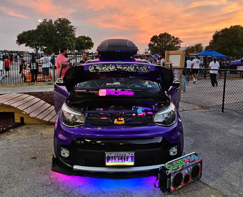 Team Hi Def A purple car with its hood open, showcasing colorful interior lights, stands out at the car show. The license plate reads "JOKSTER." In front, there's a large speaker box. As part of the JDM scene, it draws a crowd against the backdrop of a sunset sky.