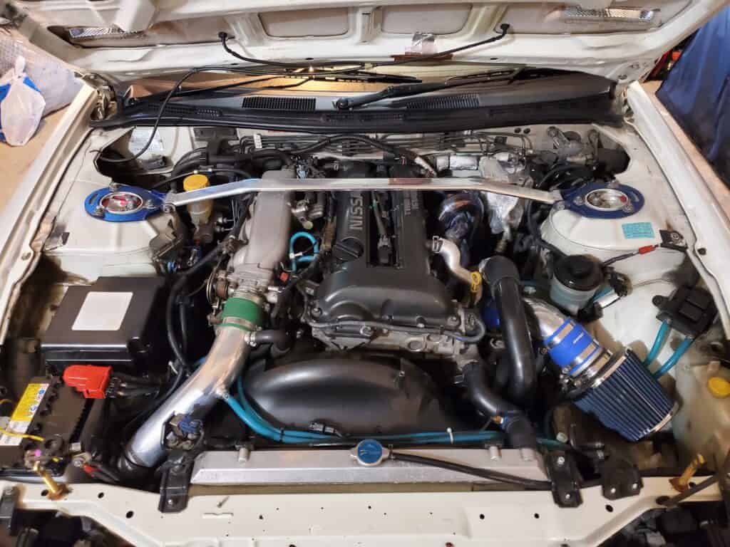 Team Hi Def The image displays the engine bay of a Nissan, showcasing its JDM-inspired setup. Visible components include a large air intake with a blue filter, strut bar, various hoses, battery, and other engine parts. The sleek engine cover proudly bears the Nissan logo and text.