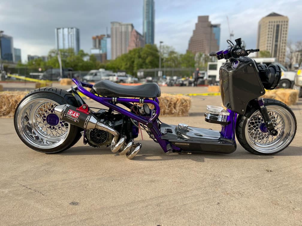 Team Hi Def A custom motorbike with a sleek frame, featuring shiny chrome wheels and exhaust, evokes the spirit of a muscle car. It has a minimalistic design with a purple and black color scheme, set against an urban backdrop with city buildings and hay bales in the distance.