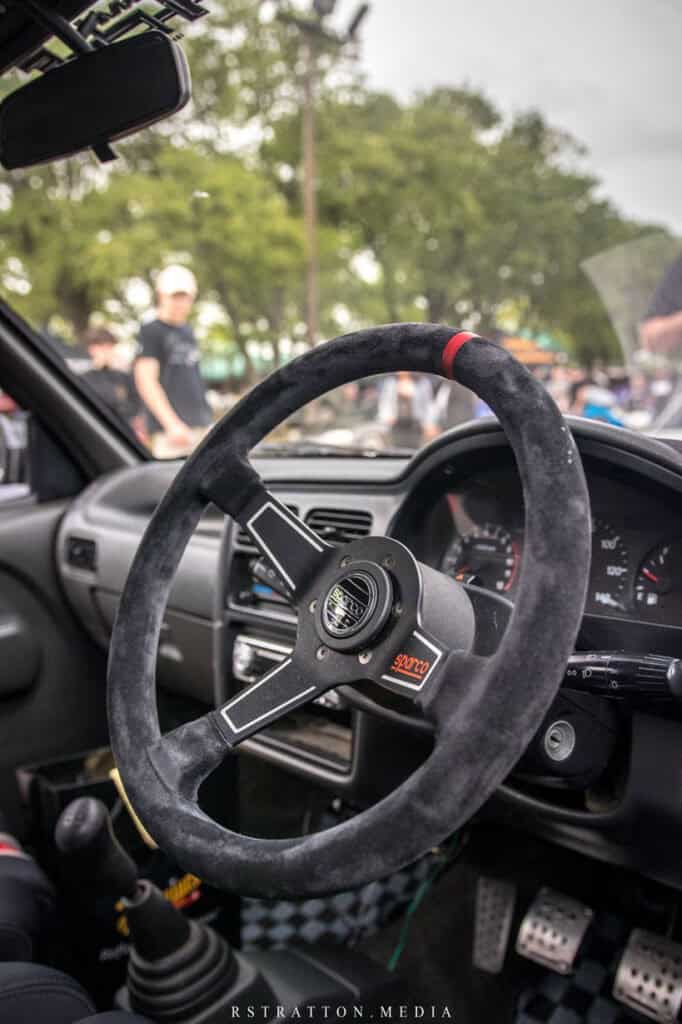 Team Hi Def The image captures the interior of an exotic car with a suede steering wheel and a dashboard featuring a tachometer. In the background, people are gathered outdoors, with trees visible under a cloudy sky.