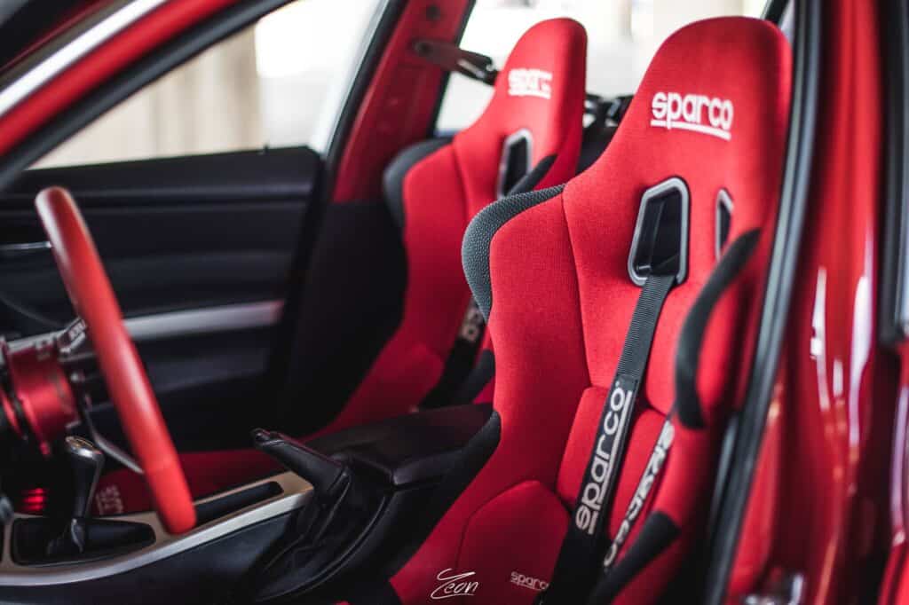 Team Hi Def The image showcases the interior of a performance car with red Sparco racing seats and a matching red steering wheel. The seats boast harness slots and black side bolsters, perfectly complementing the car's vibrant, import tuner-inspired design.