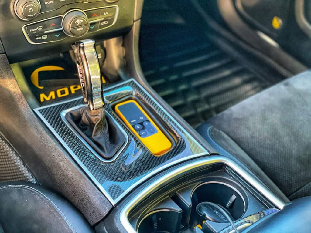 Team Hi Def Close-up of a performance car's interior featuring a gear shift and center console with carbon fiber accents. A yellow device with buttons rests in the console area, surrounded by cup holders and car controls, perfectly complementing the sleek design admired by any car club enthusiast.