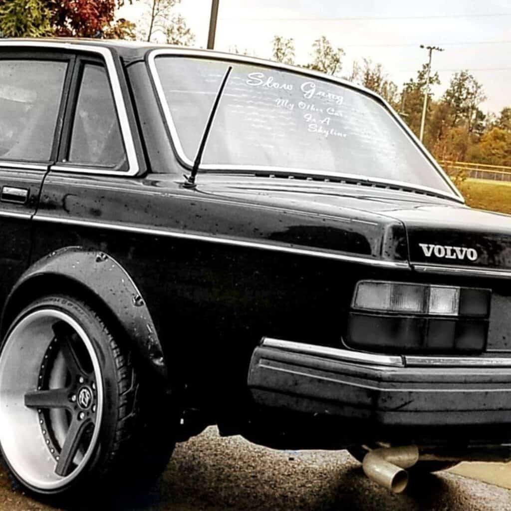 Team Hi Def Close-up of a black vintage Volvo, a true performance car, with custom wide wheels and dual exhaust pipes. The rear windshield proudly displays white text, "Show Gang... My Other Car is a Get Skyline." Behind it, trees stand beneath a cloudy sky.