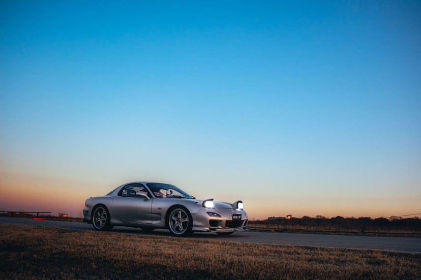 Team Hi Def A silver JDM sports car with headlights on is parked on a rural road at sunset. The sky is a gradient of blue to orange, and the landscape is flat with grass and distant trees.