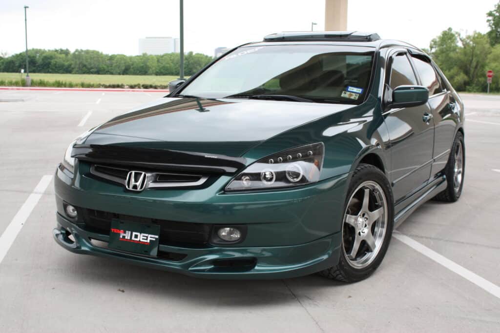 Team Hi Def A green modified Honda Accord sedan, a true JDM gem, is parked in an open lot. The performance car boasts custom alloy wheels, a black front grille, and a sporty body kit. In the background, a few trees and a distant building complete the scene.