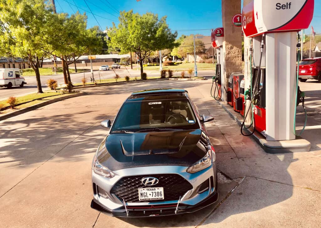 Team Hi Def A gray Hyundai, known among the local car club for its sleek design, is parked at a gas station next to a fuel pump. The scene unfolds under clear skies with trees in the background, capturing a peaceful moment in an automotive enthusiast's day.