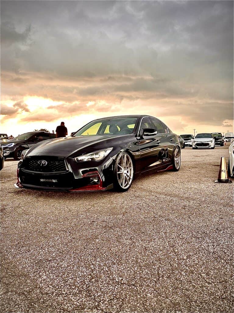 Team Hi Def A sleek black car, resembling an exotic masterpiece with polished wheels, is parked in an open area during sunset. The sky is overcast, and a few other cars are visible in the background, along with a person walking by.