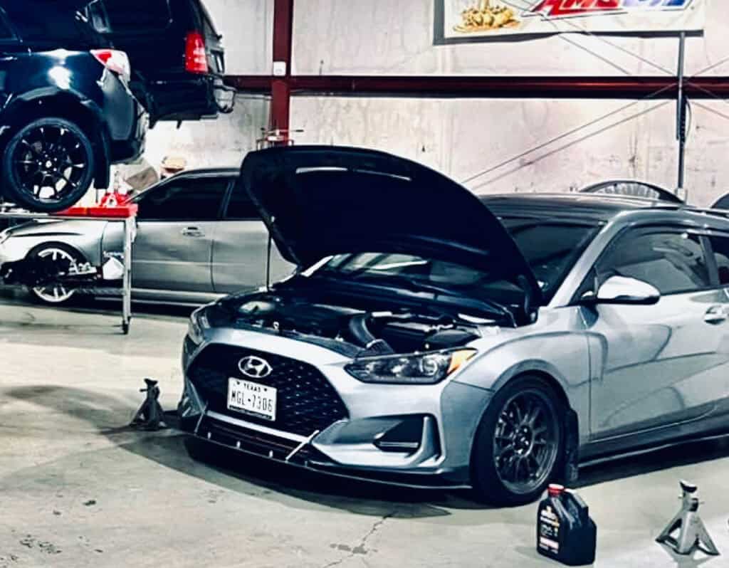 Team Hi Def A silver Hyundai, not quite a performance car but in need of some love, sits with its hood ajar on a repair platform in a bustling garage. In the backdrop, a black car is lifted high while another silver one waits patiently nearby. A bottle of motor oil rests on the floor beside them.