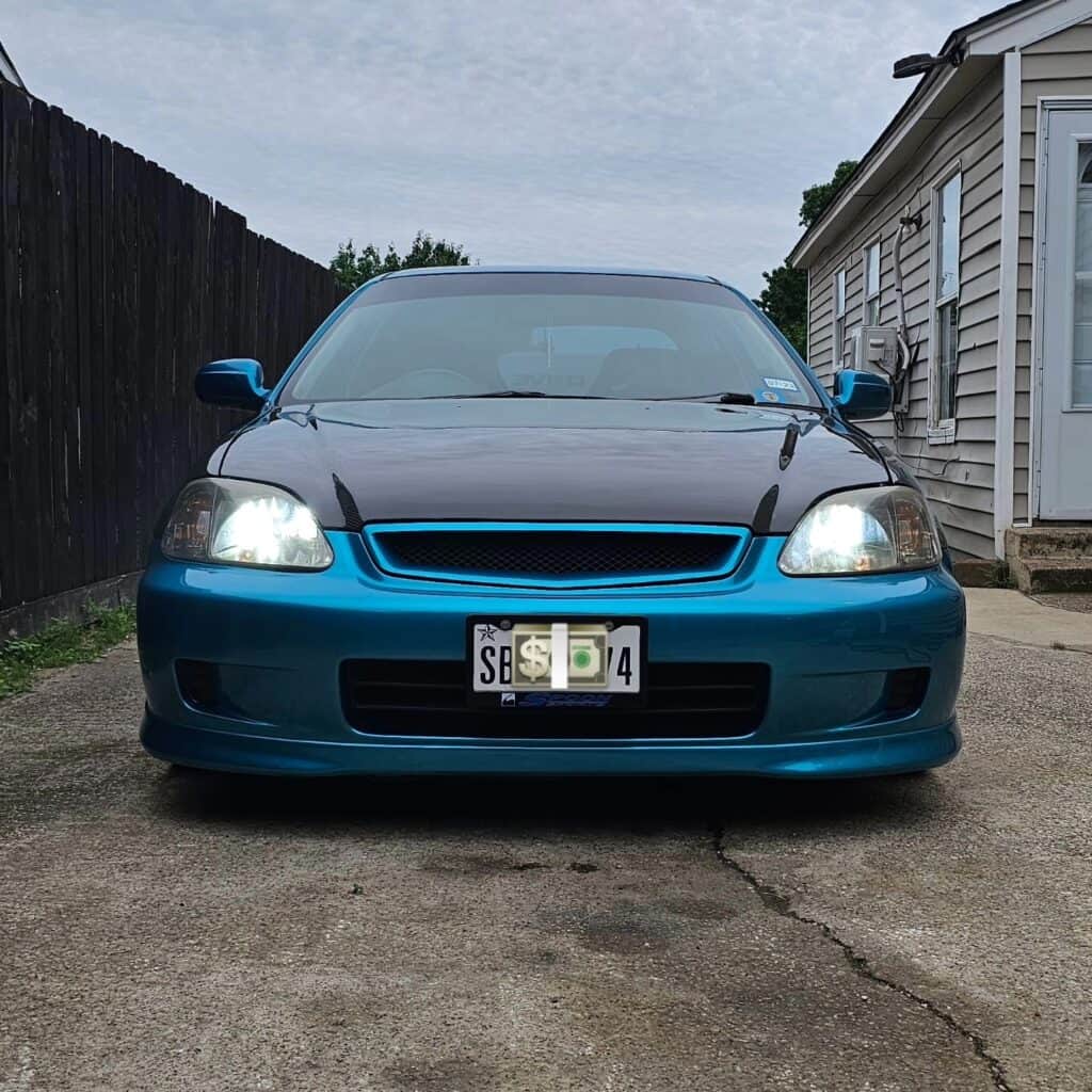 Team Hi Def A modified blue performance car is parked in a driveway. It has a black hood, and its headlights are turned on. The setting includes a wooden fence on the left and a white building on the right, perfect for impressing the local car club members.