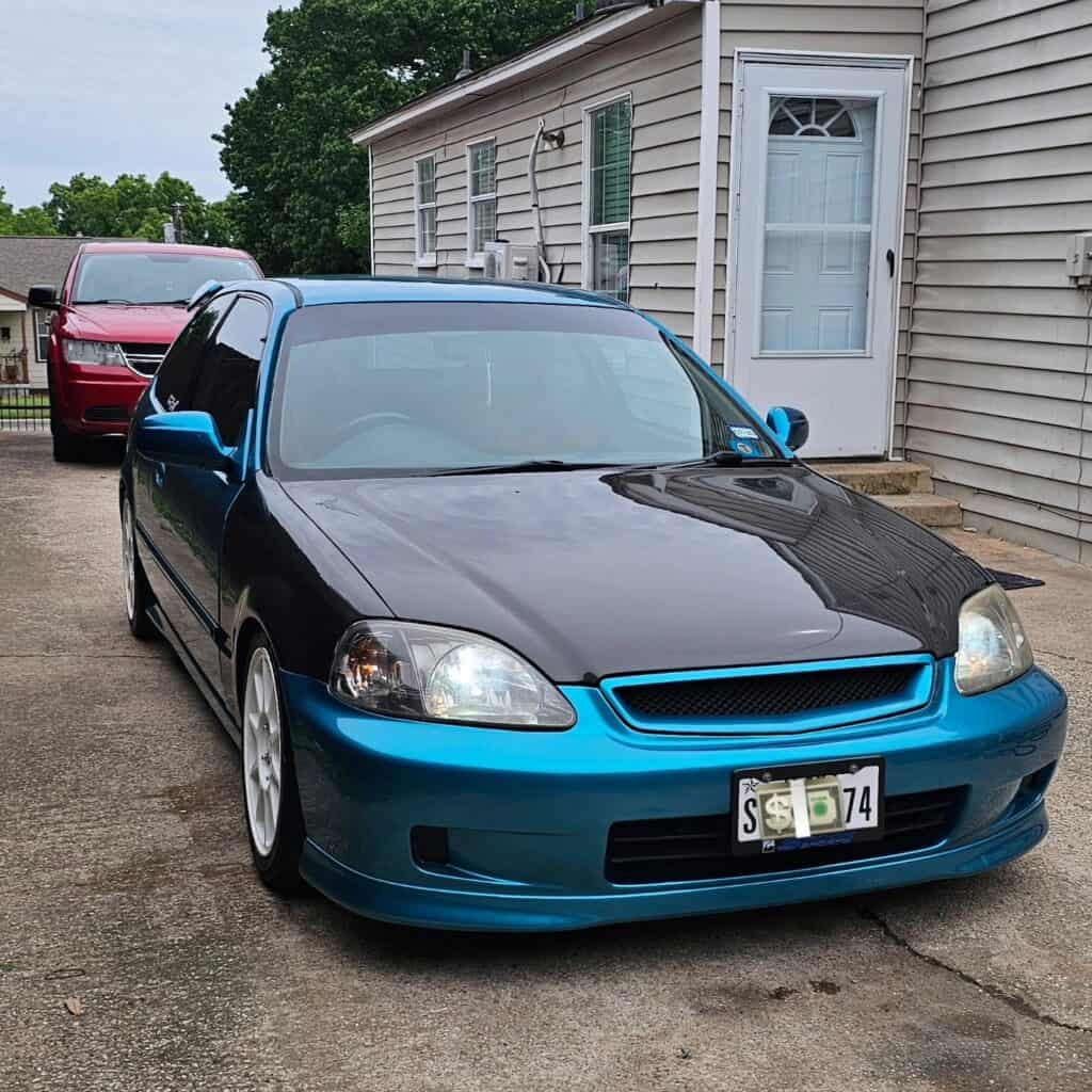 Team Hi Def A modified blue and black performance car with a custom hood and white wheels is parked in a driveway. A red vehicle, likely a fellow car club member's ride, is parked behind it, while a house with siding and a white door stands visible in the background.