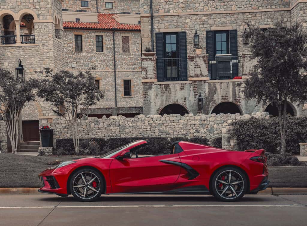 Team Hi Def A sleek red convertible sports car, a true gem among exotic cars, is parked on a street with a backdrop of a large stone building featuring arched windows and balconies. Tall trees and green shrubs line the sidewalk.