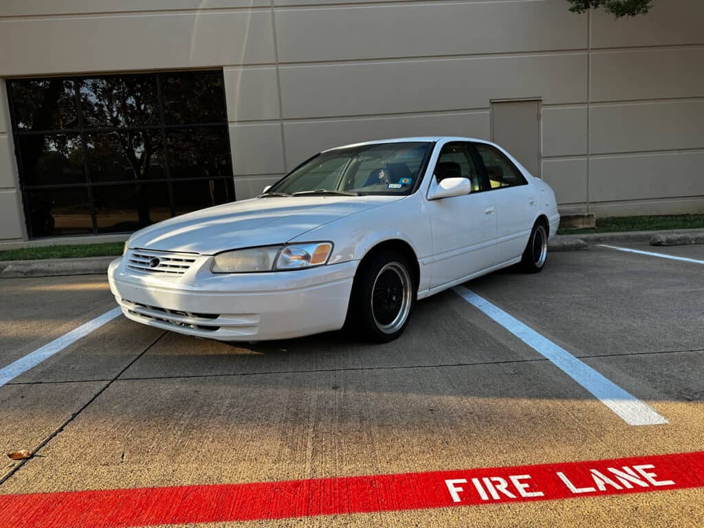 Team Hi Def A sleek white sedan, reminiscent of a JDM import tuner, is parked across a marked fire lane on a concrete surface. Positioned diagonally with front wheels slightly turned, it stands out against the backdrop of a building adorned with large windows and surrounding greenery.