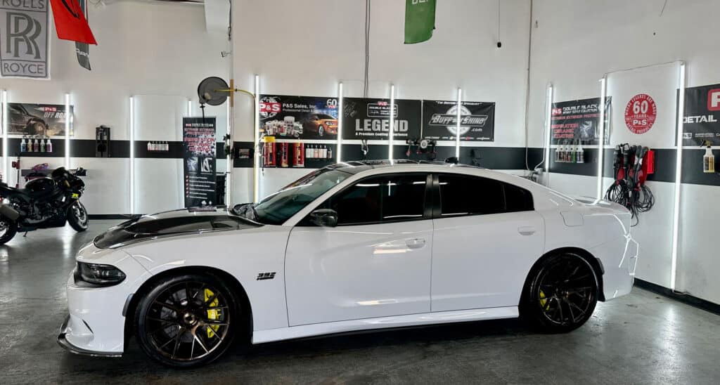 Team Hi Def A sleek white performance car with black accents and yellow brake calipers is parked indoors. The setting resembles an auto detailing garage, showcasing various car care products on shelves and walls adorned with automotive posters.