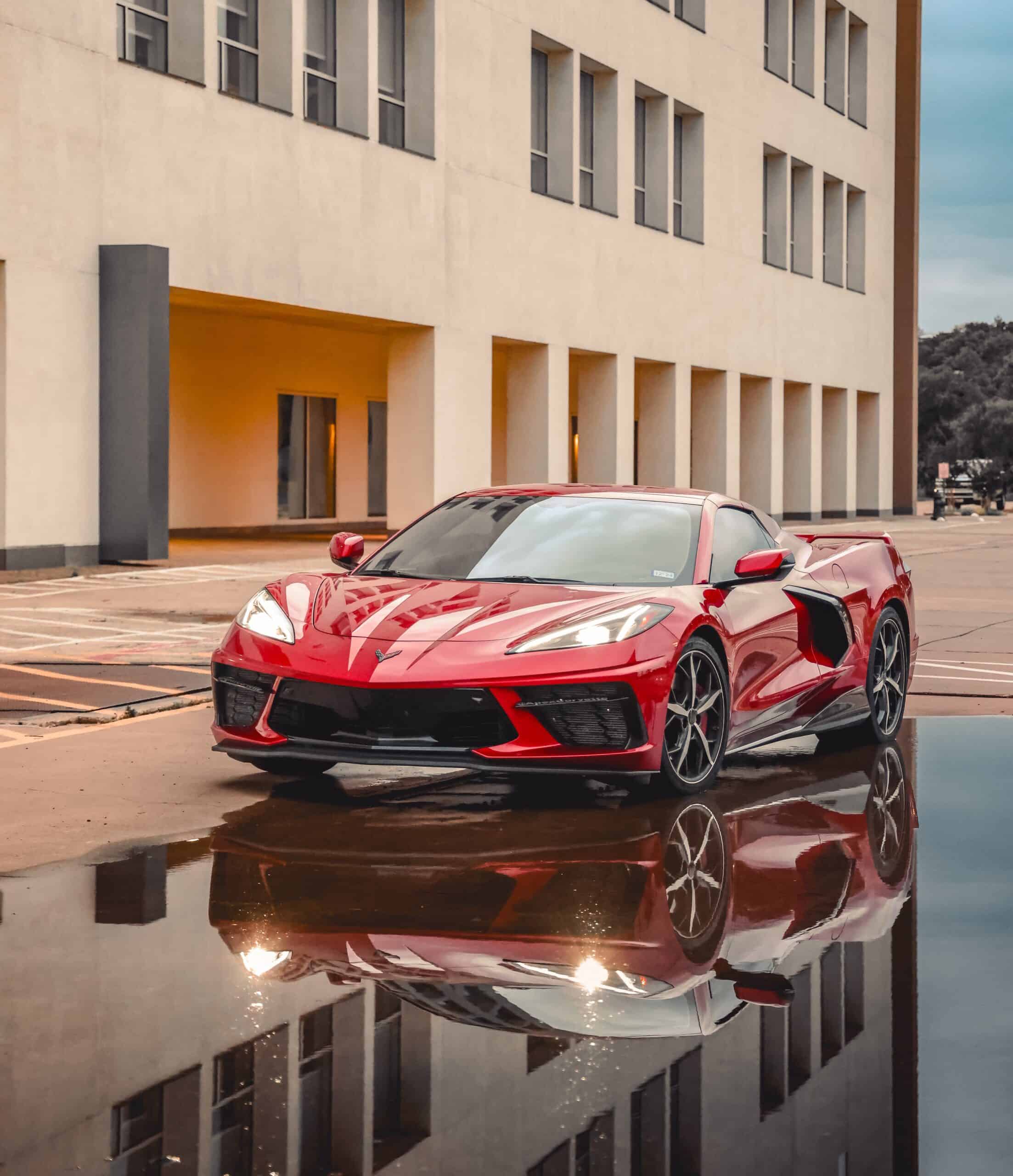 Team Hi Def A sleek, red muscle car is parked near a modern building. Its reflection shimmers in a large puddle on the ground. The background shows a clear sky and some distant trees.