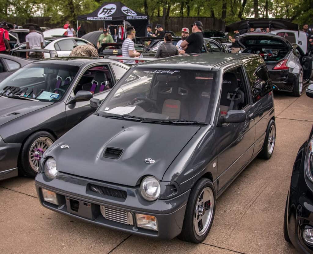 Team Hi Def A modified gray hatchback, resembling an import tuner, is parked at an outdoor car show. It features aftermarket wheels and a hood scoop. Several people are gathered around, admiring the various cars on display. Tall trees provide a scenic backdrop to the lively event.