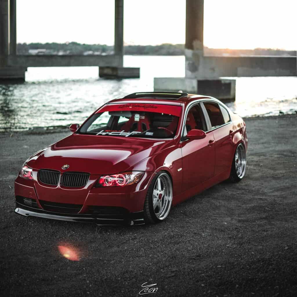 Team Hi Def A sleek, modified red BMW sedan with tinted windows and custom wheels, reminiscent of a high-end import tuner, is parked on a gravel surface near a body of water. The background features concrete pillars and a sunset sky reflecting on the water.