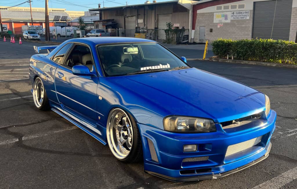 Team Hi Def A bright blue performance car is parked in an industrial area. It features a sleek, aerodynamic design with a prominent front bumper and shiny alloy wheels. A decal is visible on the windshield. In the background are buildings and greenery.