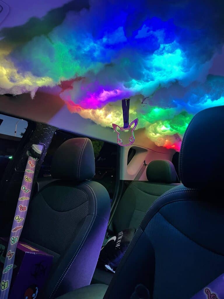 Team Hi Def The interior of an exotic car features a roof transformed into a glowing, multicolored cloud. Neon lights create a vibrant effect, while a rabbit-shaped air freshener dangles from the ceiling.