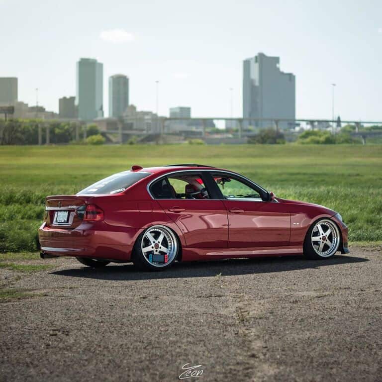 Team Hi Def A red sedan with custom rims, resembling an exotic car, is parked on a grassy field. In the background, a city skyline is visible with several tall buildings. The sky is clear and sunny.